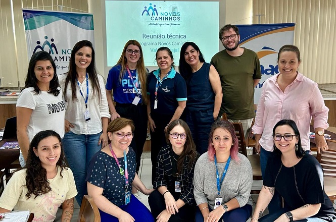A imagem mostra um grupo de pessoas posando para uma foto em uma sala. Ao fundo, há um projetor exibindo um slide com as palavras "Reunião técnica" e "Programa Novos Caminhos". Algumas pessoas estão em pé, enquanto outras estão sentadas. Eles parecem estar participando de uma reunião ou workshop. Algumas pessoas estão usando crachás. O ambiente parece ser descontraído e amigável. Há banners nas laterais da sala relacionados ao tema da apresentação.