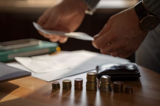Na imagem, há pilhas de moedas organizadas em uma superfície de madeira. Ao fundo, uma pessoa está manuseando papéis, possivelmente documentos ou contas. Também há uma carteira próxima às moedas. A cena sugere uma atividade ligada ao manuseio de dinheiro ou orçamento. A iluminação parece ser suave, com uma luz natural entrando na sala.