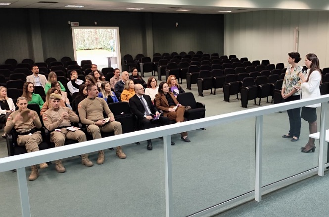 Na imagem, há uma sala de conferências com várias pessoas sentadas em poltronas, voltadas para a frente. Duas pessoas estão em pé na frente da sala, uma delas segurando o microfone, aparentemente falando ao público. Há um grupo de pessoas nas primeiras fileiras, incluindo indivíduos uniformizados, vestindo roupas formais e casuais. O ambiente é fechado, com paredes claras, piso acarpetado verde e muitas cadeiras vazias ao fundo. Parece ser um evento ou apresentação.