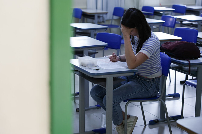 A imagem mostra uma pessoa sentada em uma sala de aula, aparentando estar concentrada em uma tarefa. Ela está usando uma camisa listrada e jeans. A sala tem várias mesas e cadeiras azuis. Além disso, há uma mochila sobre a mesa. A porta da sala está parcialmente aberta, através da qual a cena é vista.