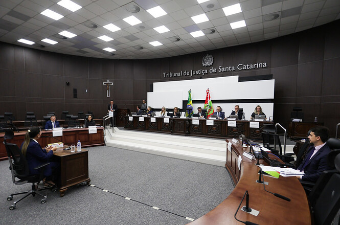 A imagem mostra uma sala de audiência no Tribunal de Justiça de Santa Catarina. O espaço é amplo e bem iluminado. Há várias pessoas sentadas em cadeiras ao redor de mesas de madeira. No fundo, há uma bancada elevada onde estão outros participantes, com bandeiras do Brasil e do estado de Santa Catarina. Na parede, está escrito "Tribunal de Justiça de Santa Catarina". A sala possui um design moderno com um teto em forma de quadrados.