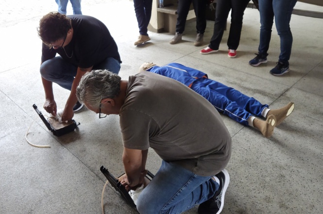 A imagem mostra um grupo de pessoas participando de um treinamento ou simulação. Duas pessoas estão ajoelhadas no chão, concentradas em atividades práticas com equipamentos em mãos. Ao fundo, uma pessoa está deitada no chão, possivelmente como parte do exercício. Várias outras pessoas estão de pé, observando atentamente o que está ocorrendo. O ambiente parece ser interno.