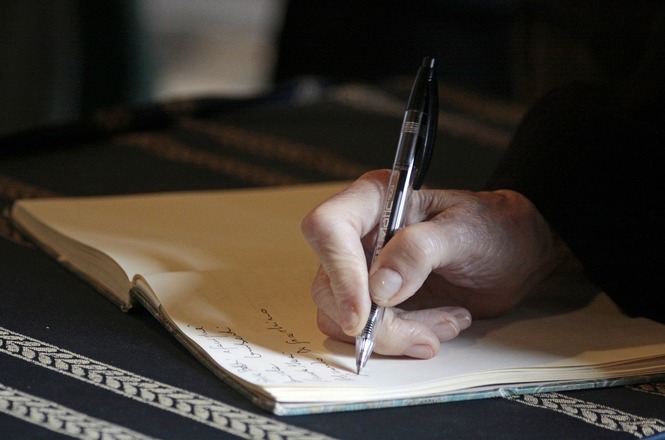 A imagem mostra uma mão segurando uma caneta enquanto escreve em um caderno, que está aberto sobre uma superfície. O foco está na ação de escrever, e o fundo é desfocado. A pessoa parece estar anotando algo com atenção. A caneta é transparente com detalhes pretos e há algumas linhas de texto já escritas na página do caderno.