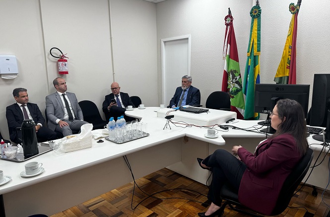 A imagem mostra uma reunião em uma sala de conferências, onde cinco pessoas estão sentadas ao redor de uma mesa em formato de "U". Há duas bandeiras na parede, junto com um extintor de incêndio. Na mesa, há xícaras de café, garrafas de água, e um monitor de computador. As pessoas estão vestidas formalmente, com ternos e roupas de trabalho. A sala parece ser um ambiente profissional.