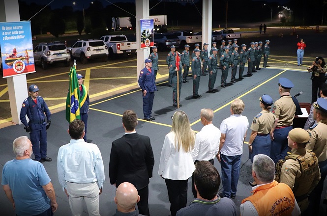 A imagem mostra uma cerimônia oficial com a participação de oficiais militares. No centro, há uma bandeira do Brasil sendo segurada por um dos oficiais. Várias pessoas estão de pé, voltadas para a bandeira, incluindo civis e outros militares em uniforme. No fundo, há uma linha de oficiais em posição de sentido. O local parece ser uma área coberta, possivelmente um pátio. Placas e veículos estão visíveis ao fundo do lado de fora.
