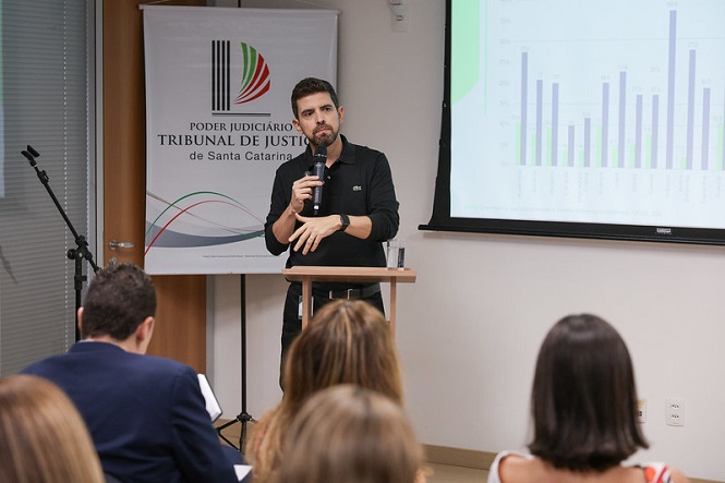 Na imagem, há um homem segurando um microfone e falando em frente a um pequeno púlpito. Ele está em uma apresentação com um público à sua frente, aparentemente apresentando dados mostrados em um gráfico projetado em uma tela à direita. Ao fundo, há um banner com o logotipo e o nome "Tribunal de Justiça de Santa Catarina". O ambiente parece ser uma sala de conferências ou auditório.