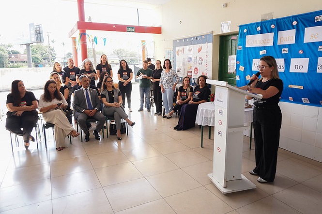 A imagem mostra uma mulher em pé ao lado de um púlpito, falando para um grupo de pessoas sentadas. O ambiente parece ser uma sala ou auditório, com um mosaico colorido e cartazes nas paredes. Há frases como "Conhecer para se..." e "Vivências no..." em um mural azul. Algumas pessoas na plateia usam roupas formais e outras estão em trajes mais casuais. Ao fundo, há uma fila de pessoas em pé, possivelmente participando de um evento ou cerimônia. A área tem uma aparência organizada e bem iluminada.