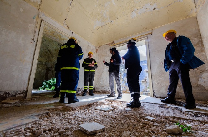 A imagem mostra cinco pessoas reunidas em um local que parece ser uma construção antiga e desgastada. Eles estão vestindo roupas de trabalho, incluindo capacetes amarelos e uniformes. O chão está coberto de destroços e a estrutura do local parece estar danificada. Eles aparentam estar discutindo ou avaliando a situação do ambiente ao redor. A luz entra por uma abertura que pode ser de uma porta ou janela, iluminando a cena.