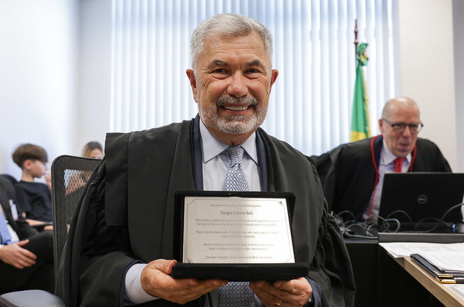 Na imagem, há um homem sorridente, vestindo uma toga, segurando uma placa de homenagem. Atrás dele, há outras pessoas, incluindo um homem que parece estar trabalhando em um laptop. Também é visível uma bandeira do Brasil ao fundo. A cena sugere um ambiente formal, possivelmente relacionado a uma cerimônia ou evento oficial.