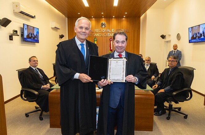 A imagem mostra homens vestidos com togas em um ambiente formal, possivelmente uma cerimônia em um tribunal. Dois deles estão em destaque na frente, segurando um documento enquadrado. Ao fundo, há outras pessoas sentadas em cadeiras atrás de uma bancada. Na parede, lê-se "Regional Eleitoral de Santa Catarina". O ambiente parece ser uma sala de audiências ou tribunal, com iluminação de teto e monitores exibindo imagens da mesma sala.
