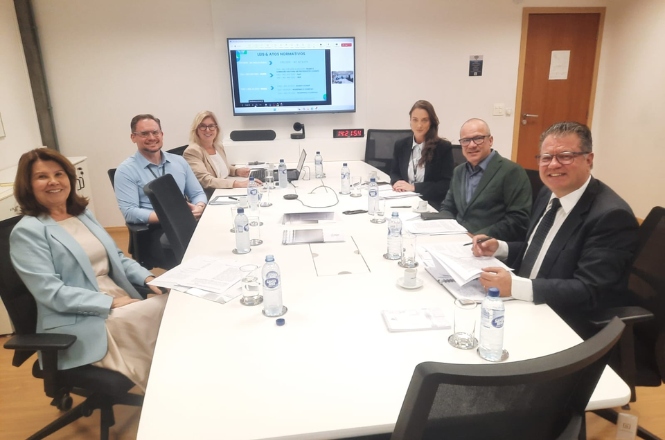 A imagem mostra um grupo de sete pessoas sentadas ao redor de uma mesa de reunião em uma sala de conferências. A mesa está coberta com garrafas de água, copos, documentos e dispositivos eletrônicos. Na parede ao fundo, há uma tela exibindo uma apresentação. Todos parecem estar envolvidos em uma discussão ou reunião de negócios. A sala é bem iluminada e há um ambiente formal.