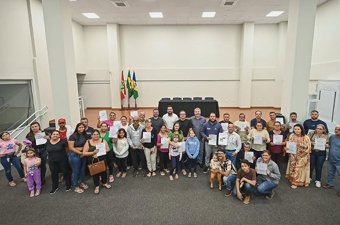 A imagem mostra um grupo grande de pessoas posando juntas em um ambiente interno, aparentemente em uma espécie de auditório ou salão de eventos. Muitas das pessoas estão segurando certificados ou documentos e há um ar de celebração ou comemoração. No fundo, vê-se uma mesa coberta com uma toalha preta e bandeiras ao lado, sugerindo um evento formal ou solene. Algumas crianças fazem parte do grupo, e todos parecem estar sorridentes.