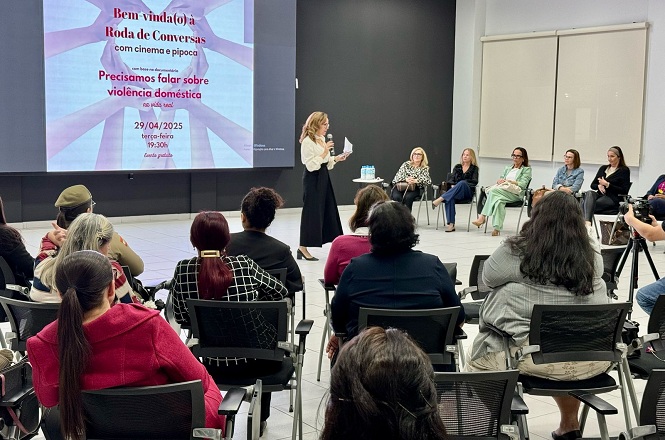 A imagem mostra um evento denominado "Roda de Conversas", cujo tema é "Precisamos falar sobre violência doméstica". Uma mulher está de pé, falando ao microfone, enquanto outras pessoas estão sentadas ouvindo. O local tem um ambiente fechado com cadeiras e decoração simples. O evento está programado para o dia 29/04/2025, das 19h às 20h30.