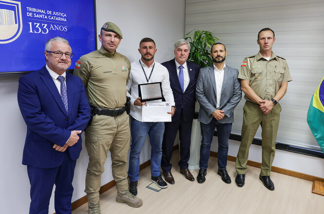 Na imagem, há seis homens posando para uma foto em um ambiente interno. Eles estão de pé lado a lado. Dois dos homens estão vestindo uniformes oficiais. Um homem ao centro segura um certificado e uma placa. No fundo, à esquerda, há uma tela exibindo "Tribunal de Justiça de Santa Catarina 133 anos". À direita, é visível parte de uma bandeira do Brasil. As pessoas estão posicionadas de forma formal, possivelmente para uma ocasião especial ou cerimônia.