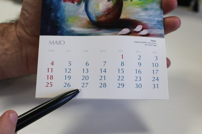 A imagem mostra um calendário do mês de maio. As semanas começam no domingo e os dias estão numerados de 1 a 31. A data 1º de maio está em vermelho, assim como os domingos. Um dedo segura o calendário enquanto uma caneta está apontando para o dia 27. Acima do calendário, há uma imagem colorida de um vaso com flores.