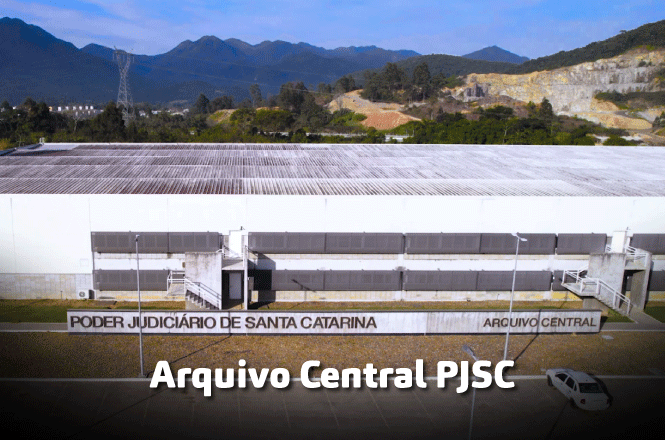 A imagem mostra um edifício do "Poder Judiciário de Santa Catarina", identificado como "Arquivo Central PJSC". O prédio é grande e moderno, com uma fachada branca e detalhes em concreto. Está situado em uma área cercada de montanhas e vegetação, sob um céu azul. Na parte inferior da imagem, há um texto destacado que diz "Arquivo Central PJSC".