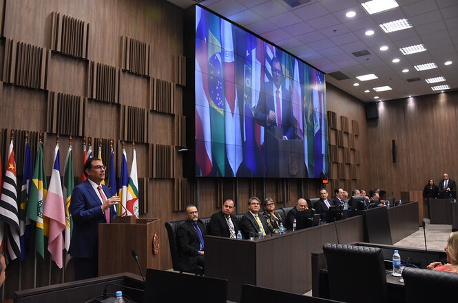 Foto horizontal: Francisco Oliveira Neto em pé no púlpito discursa em plenário, autoridades à mesa e telão ao fundo.