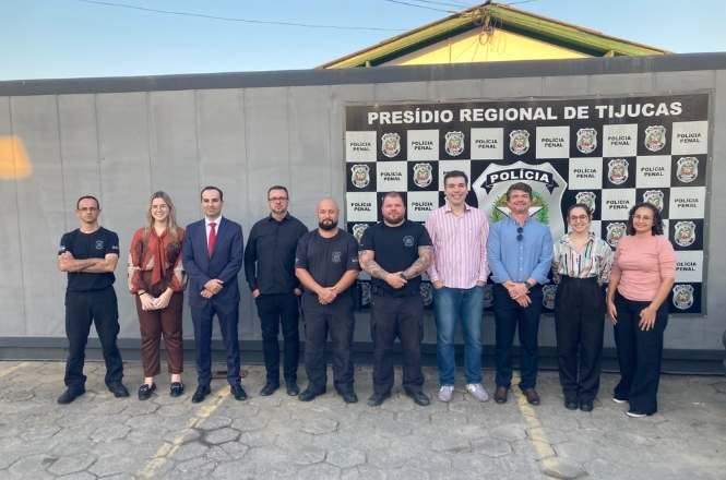 A imagem mostra um grupo de pessoas posando para uma foto em frente a uma parede com o logotipo da "Polícia Penal" do "Presídio Regional de Tijucas". Eles estão em pé, lado a lado. A parede atrás deles tem várias placas, e eles estão em uma área externa com piso de paralelepípedo. Ao fundo, há um pequeno prédio com um telhado de duas águas.