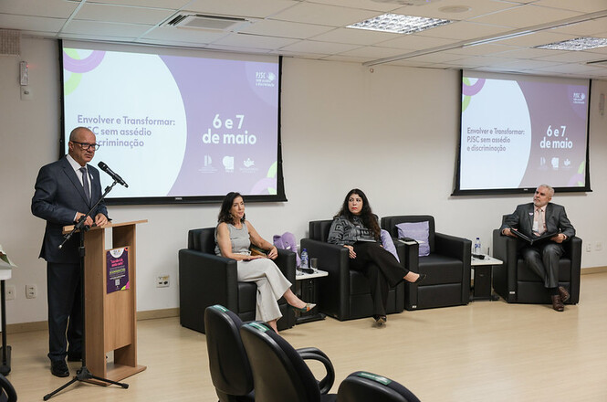 A imagem mostra um evento em um auditório. Há quatro pessoas na foto: uma pessoa de pé à esquerda, possivelmente discursando em um púlpito, e três pessoas sentadas em poltronas à direita. No fundo, há dois telões exibindo a mensagem "Envolver e Transformar: PSSC sem assédio e discriminação", com as datas "6 e 7 de maio". O ambiente é profissional, e todos estão vestidos formalmente. O cenário sugere uma palestra ou conferência sobre tópicos relacionados a assédio e discriminação no ambiente de trabalho.