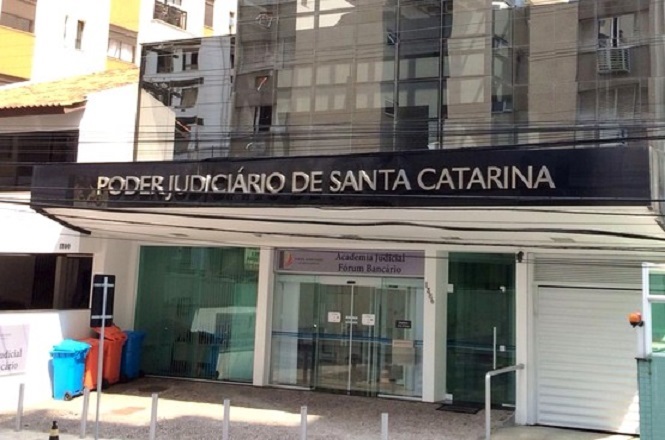 A imagem mostra a fachada de um edifício com a placa "Poder Judiciário de Santa Catarina". Abaixo, há outra placa indicando "Academia Judicial" e "Fórum Bancário". Na frente do edifício, há uma entrada de vidro e algumas lixeiras coloridas à esquerda. A arquitetura é moderna, com linhas retas e vidro.
