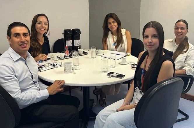 Na imagem, há cinco pessoas sentadas ao redor de uma mesa redonda, participando de uma reunião. A mesa está equipada com copos, xícaras e garrafas térmicas. Todos na imagem estão sorrindo e parecem estar em um ambiente profissional ou de escritório. A sala tem paredes claras e uma atmosfera de colaboração é aparente.