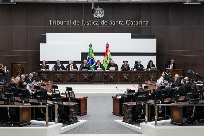 A imagem mostra uma sessão no Tribunal de Justiça de Santa Catarina. Há várias pessoas sentadas em cadeiras dispostas em semicírculo, com uma bancada central onde estão localizados os juízes e autoridades. Na parede ao fundo, está escrito “Tribunal de Justiça de Santa Catarina”. Também há bandeiras do Brasil e do estado de Santa Catarina posicionadas atrás dos juízes. O ambiente parece ser formal com um design interior elegante.