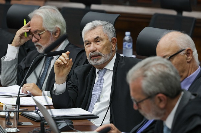 A imagem mostra quatro homens sentados em uma mesa, provavelmente em um ambiente formal como uma reunião ou sessão de tribunal. Eles estão vestidos com ternos e togas. O homem ao centro parece estar falando ou fazendo uma declaração, enquanto os outros estão ouvindo atentamente e fazendo anotações. Há microfones diante deles, sugerindo que estão participando de uma discussão oficial. No fundo, podem ser vistos cadeiras e uma garrafa de água sobre a mesa.