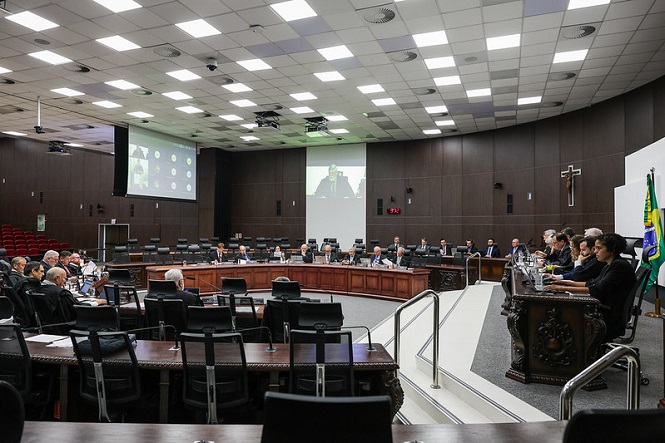 A imagem mostra uma sala ampla, que parece ser de um tribunal ou espaço governamental. Há várias pessoas sentadas em semicírculo ao redor de uma mesa central. Na frente, há um púlpito com câmeras e microfones, e algumas bandeiras no canto direito, incluindo a bandeira do Brasil. Há um grande telão de projeção ao fundo, que exibe uma imagem no estilo de videoconferência. As paredes são de madeira escura, e o teto é coberto por painéis brancos com luminárias embutidas. Também há um crucifixo na parede ao fundo.
