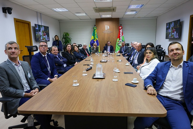 A imagem mostra uma reunião em uma sala de conferências. Várias pessoas estão sentadas ao redor de uma mesa retangular. A sala é bem iluminada e há bandeiras do Brasil e de Santa Catarina ao fundo. Algumas pessoas estão vestidas formalmente, usando ternos e roupas sociais. Há itens como copos e garrafas sobre a mesa, e algumas pessoas estão olhando para a câmera. O ambiente parece ser profissional.
