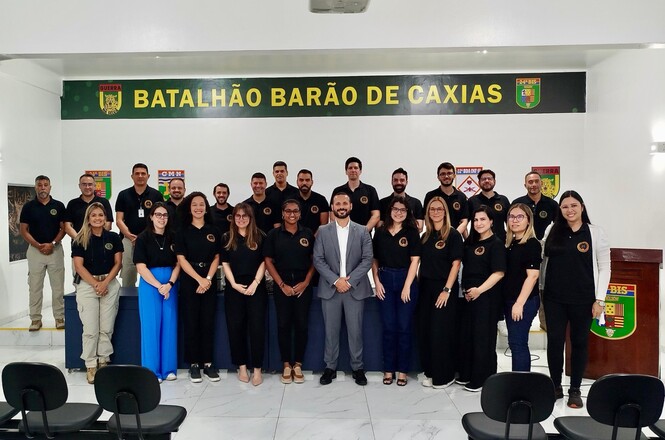A imagem mostra um grupo de pessoas posando para uma foto em um ambiente fechado. Eles estão organizados em duas fileiras, alguns em pé na parte de trás e outros agachados ou em pé na frente. A maioria veste camisetas pretas com um distintivo no peito, enquanto uma pessoa no centro está vestindo um terno cinza. Ao fundo, há uma parede com o texto "BATALHÃO BARÃO DE CAXIAS" escrito em um painel acima. O ambiente parece ser uma sala de reuniões ou auditório, com cadeiras vazias em primeiro plano.