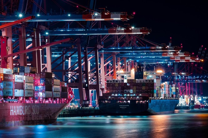 A imagem mostra um porto à noite, iluminado por luzes artificiais. No porto, há grandes guindastes carregando e descarregando contêineres de navios de carga. Os navios estão ancorados ao longo de um cais, e suas luzes refletem na água. A cena destaca a atividade industrial e a infraestrutura do porto.