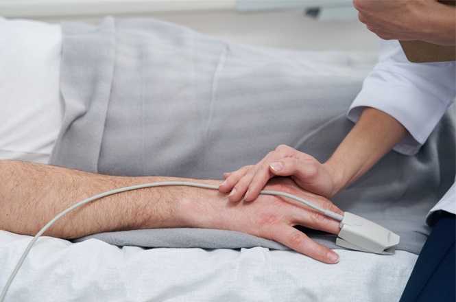 A imagem mostra a mão de uma pessoa deitada em uma cama, coberta com um cobertor cinza. A pessoa está com um oxímetro no dedo, conectado a um fio. Outra pessoa está segurando a mão da pessoa deitada, possivelmente fornecendo cuidados médicos ou apoio.
