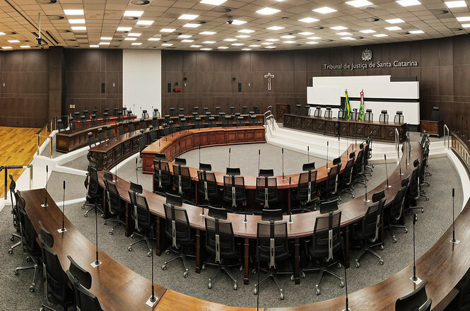 A imagem retrata uma sala de tribunal moderna e espaçosa, provavelmente do Tribunal de Justiça de Santa Catarina, conforme indicado na parede ao fundo. O espaço é bem iluminado, com teto repleto de luzes embutidas. Há várias cadeiras pretas organizadas em torno de mesas marrons em formato semicircular. No centro, há uma área elevada com mais cadeiras, possivelmente para juízes ou autoridades do tribunal. Na parede ao fundo, encontram-se bandeiras e um crucifixo à direita.