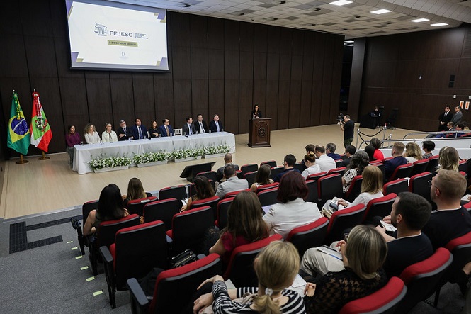 A imagem mostra um auditório com várias pessoas sentadas. No palco, há uma mesa com diversas pessoas sentadas, possivelmente autoridades ou palestrantes. Há duas bandeiras, do Brasil e de Santa Catarina, posicionadas à esquerda do palco. Uma pessoa está em pé, aparentemente falando em um púlpito. O público está atento, e o ambiente é formal, sugerindo um evento oficial ou uma conferência.