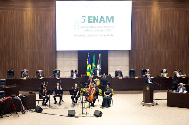 A imagem mostra uma cerimônia formal em um grande auditório. No palco, há várias pessoas sentadas em cadeiras alinhadas. Há um grupo de músicos com violinos, um violoncelo e uma cantora no centro, prontos para uma apresentação. No fundo, bandeiras estão dispostas atrás das pessoas sentadas no palco. Acima, um grande painel exibe "5º ENAM Encontro Nacional de Memória do Poder Judiciário 2023 Memória, Cultura e Diversidade". Há também um púlpito à direita, onde uma pessoa parece estar falando ao microfone. O ambiente é elegante e bem iluminado.