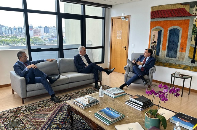 Foto horizontal: Francisco Oliveira Neto gesticula em reunião informal, autoridades sentadas, ambiente interno, tons claros.