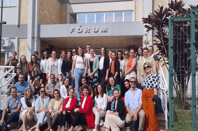 A imagem mostra um grupo grande de pessoas posando para uma foto em frente a um edifício com a placa "FÓRUM" visível ao fundo. As pessoas estão sorrindo e parecem estar participando de uma atividade ou evento conjunto. Algumas pessoas estão sentadas nos degraus da entrada, enquanto outras estão em pé. A atmosfera é de camaradagem e coletividade. Há também vegetação e uma cerca ao lado direito da imagem.