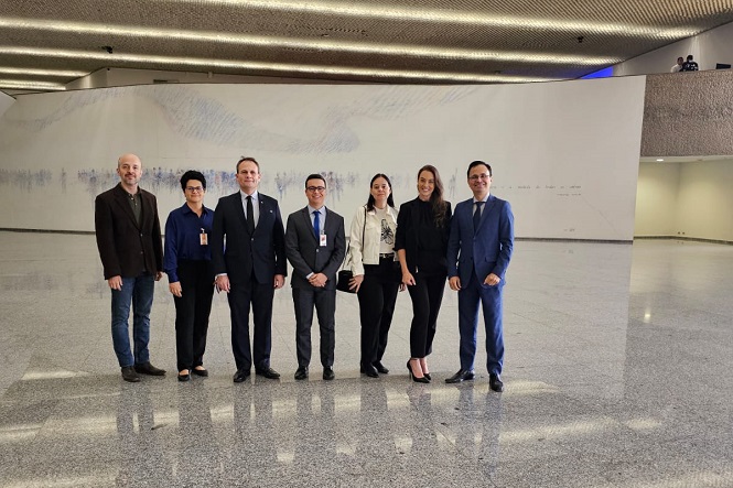 A imagem mostra um grupo de sete pessoas posando para uma foto em um ambiente interno. O chão é de mármore ou granito, e o teto tem um design moderno com iluminação embutida. As pessoas estão vestidas de forma formal, como se estivessem em um evento ou reunião de negócios. No fundo, há uma grande parede clara com uma obra de arte abstrata em tons suaves.