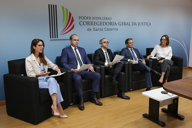 Na imagem, há cinco pessoas sentadas em cadeiras pretas em uma sala. Elas estão usando trajes formais e segurando papéis. Atrás delas, há uma parede com o logotipo e o nome "Poder Judiciário Corregedoria-Geral da Justiça de Santa Catarina". A atmosfera parece ser de um ambiente formal ou reunião. A sala tem piso de madeira e uma mesa à direita.