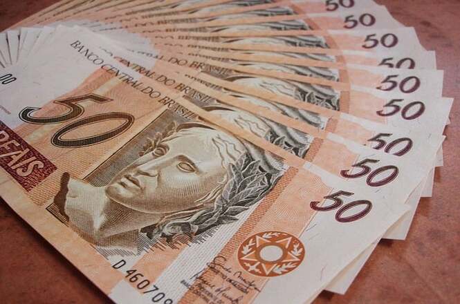 Na imagem, há várias notas de cinquenta reais dispostas em formato de leque. As notas possuem a inscrição "Banco Central do Brasil" e apresentam a figura de uma representação alegórica da República. A cor predominante é um tom alaranjado.