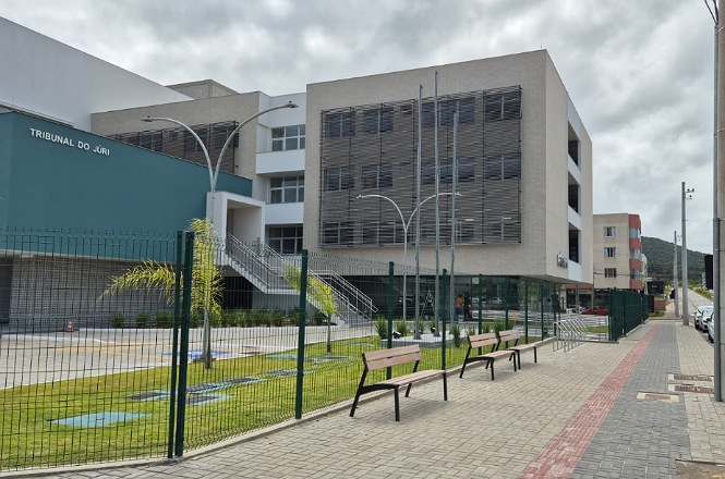 A imagem mostra um edifício moderno, identificado como "Tribunal do Júri". A estrutura tem varandas e grandes janelas. Na frente, há um jardim bem cuidado com algumas plantas e bancos para sentar. O prédio é cercado por uma grade, e a calçada ao lado é pavimentada e possui postes de luz. O céu está nublado.