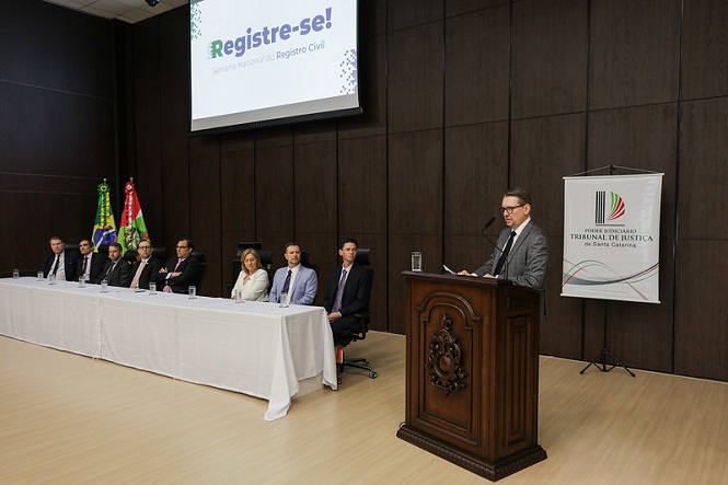 A imagem mostra um evento formal em um auditório. Há uma mesa comprida com várias pessoas sentadas, vestidas de terno e gravata, exceto uma mulher de casaco claro. À direita, há um homem em pé, falando em um púlpito de madeira escura. No fundo, há duas bandeiras e um banner do "Poder Judiciário Tribunal de Justiça de Santa Catarina". No telão, lê-se "Registre-se! Selo Nacional da Registro Civil".