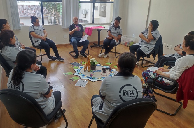A imagem mostra um grupo de pessoas sentadas em cadeiras dispostas em círculo em uma sala. Elas estão participando de uma reunião ou atividade em grupo. No centro do círculo, há um tapete com objetos, como flores e cartões distribuídos. As pessoas parecem estar prestando atenção ou discutindo algo. Algumas estão usando uniformes com o logotipo da "BRA". As janelas ajustam a iluminação natural do ambiente.