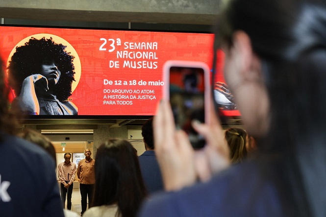 A imagem mostra um grupo de pessoas reunidas em frente a uma grande tela eletrônica que exibe um cartaz. No cartaz, está escrito "23ª Semana Nacional de Museus", que ocorrerá de 12 a 18 de maio, com o tema "Do arquivo ao átomo: a história da justiça para todos". Uma pessoa está tirando uma foto da tela com o celular. A imagem no cartaz apresenta uma figura artística. A cena parece ser em um ambiente interno, possivelmente em um evento ou conferência.