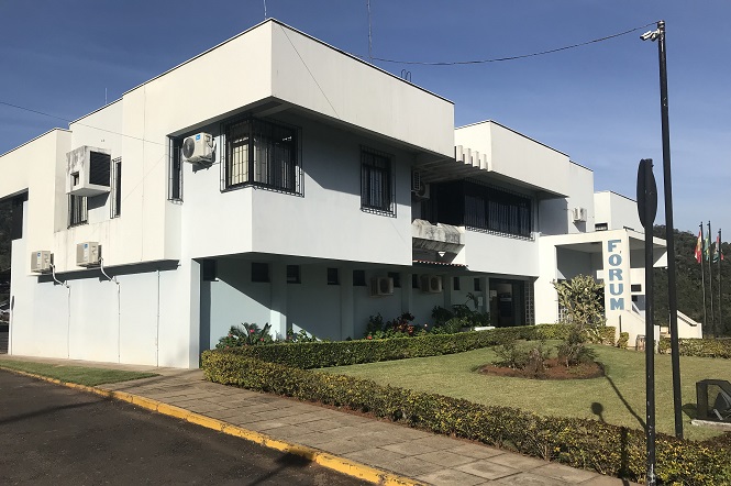 A imagem mostra um edifício moderno de cor branca com dois andares. Na fachada, há placas de ar-condicionado visíveis nas janelas. A frente do prédio possui um jardim bem cuidado com gramado e arbustos. Na lateral do edifício, há uma placa vertical com a palavra "FÓRUM". O céu está claro e ensolarado, o que sugere que a foto foi tirada durante o dia. No canto direito da imagem, há bandeiras hasteadas.