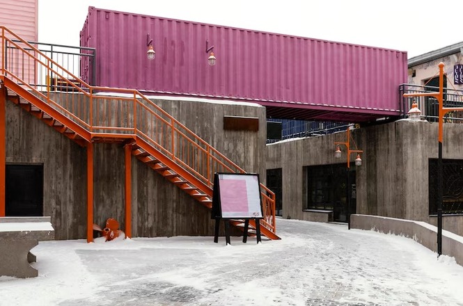 A imagem mostra uma cena urbana em um ambiente coberto de neve. No centro, há uma escada de metal laranja que leva a uma estrutura de concreto. Acima desta estrutura, um contêiner rosa está posicionado horizontalmente, atuando como uma espécie de passarela ou ligação entre edifícios. Perto da base da escada, há um pequeno hidrante de incêndio também pintado de laranja. No chão, pode-se ver a neve espalhada. As cores vivas do contêiner e da escada contrastam com o cinza do concreto e o branco da neve. Uma placa retangular com um suporte preto está colocada ao lado da escada.
