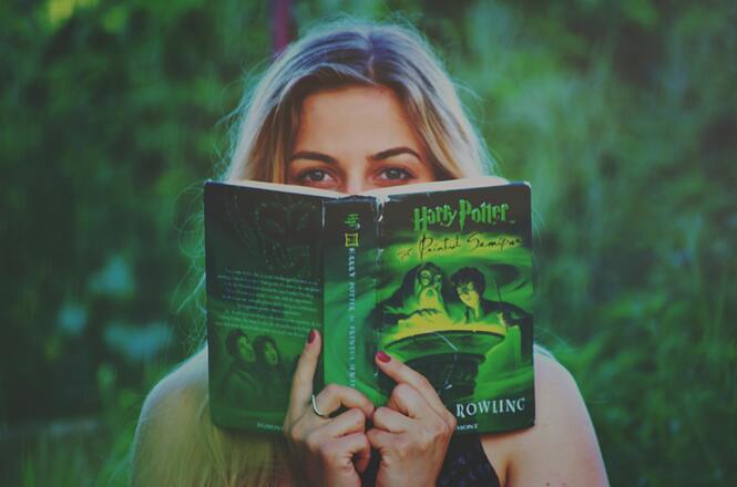 A imagem mostra uma pessoa segurando um livro, com o título "Harry Potter e o Enigma do Príncipe" visível na capa. A capa do livro é verde e apresenta personagens em um cenário iluminado por uma luz mágica. A pessoa está parcialmente escondida atrás do livro, com o olhar direcionado para frente. O fundo é desfocado, sugerindo um ambiente ao ar livre com vegetação.
