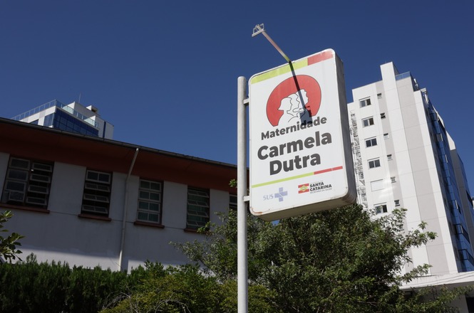 A imagem mostra a placa de uma maternidade chamada "Carmela Dutra". A placa é colorida, com um fundo vermelho e um desenho estilizado em branco. Abaixo do nome, há menções ao SUS e ao Governo de Santa Catarina. Ao fundo, aparecem edifícios e vegetação sob um céu azul.