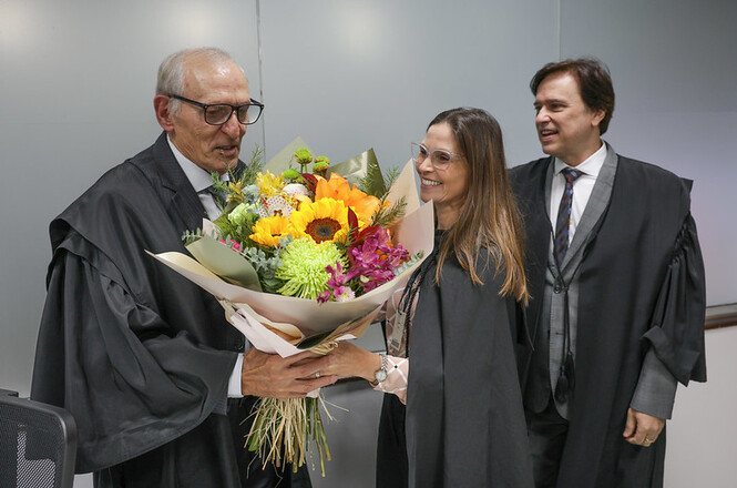 Na imagem, há três pessoas vestindo togas formais. A pessoa à esquerda está segurando um buquê de flores coloridas e parece estar sendo presenteado(a) pela pessoa ao centro, que está sorrindo. A terceira pessoa, à direita, também está sorrindo enquanto observa a cena. Todos estão em um ambiente interno, provavelmente durante algum evento formal ou celebração.