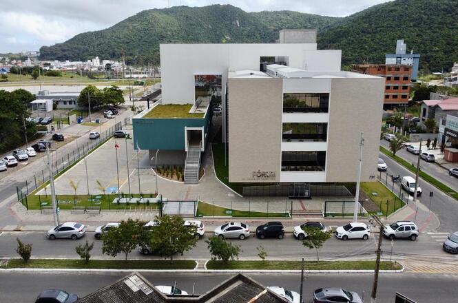 A imagem mostra um edifício moderno, que parece ser um prédio público com placas de identificação na frente, possivelmente um fórum ou uma sede administrativa. O prédio tem um design arquitetônico contemporâneo, com linhas retas e janelas grandes. Há uma área verde com árvores e bandeiras na frente, além de um estacionamento com vários carros. Ao fundo, é possível ver colinas cobertas de vegetação, e o clima parece nublado. A rua em frente ao edifício tem movimento de veículos.